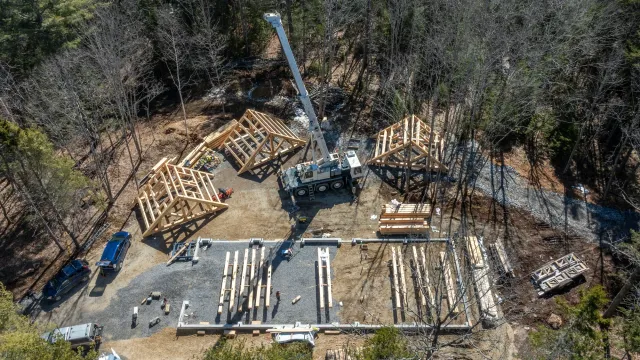 Aerial view of a house construction site in a wooded area with a crane lifting wooden roof trusses and construction vehicles parked nearby.