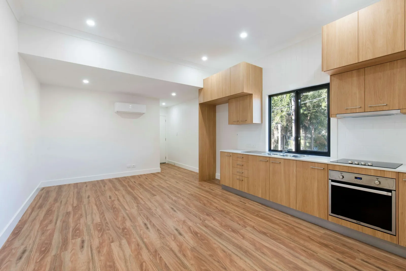 Empty modern kitchen with wooden cabinets, hardwood floor, large window, and white walls.