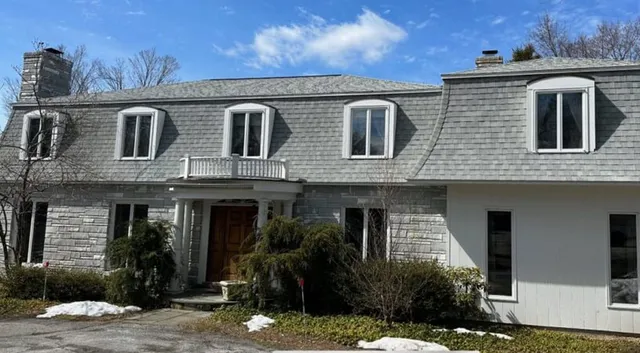 Chris Battaini Roofing - A large gray stone house with a wooden door, dormer windows, and a snowy front yard under a partly cloudy blue sky.