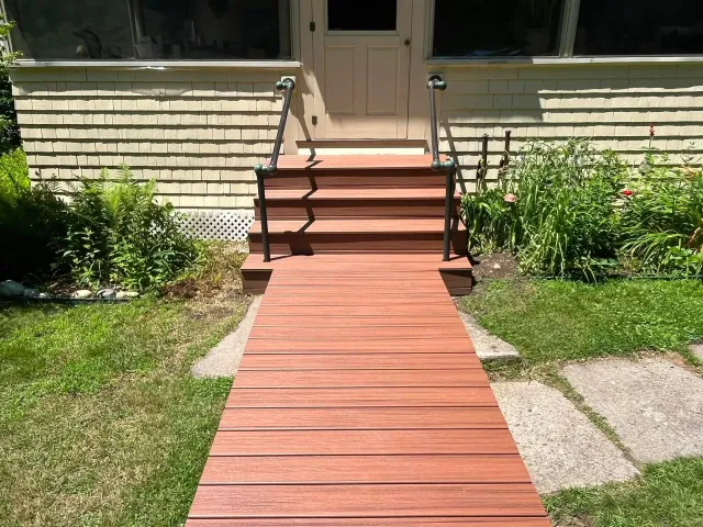 Front walkway with wooden steps leading to a tan house entrance, bordered by grass and shrubs.