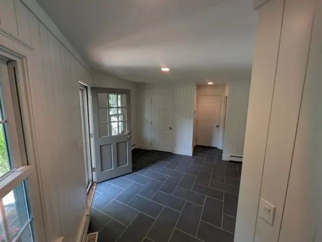 Empty room with white paneled walls, black tiled floor, windows, a door with glass panes, and recessed ceiling lights.