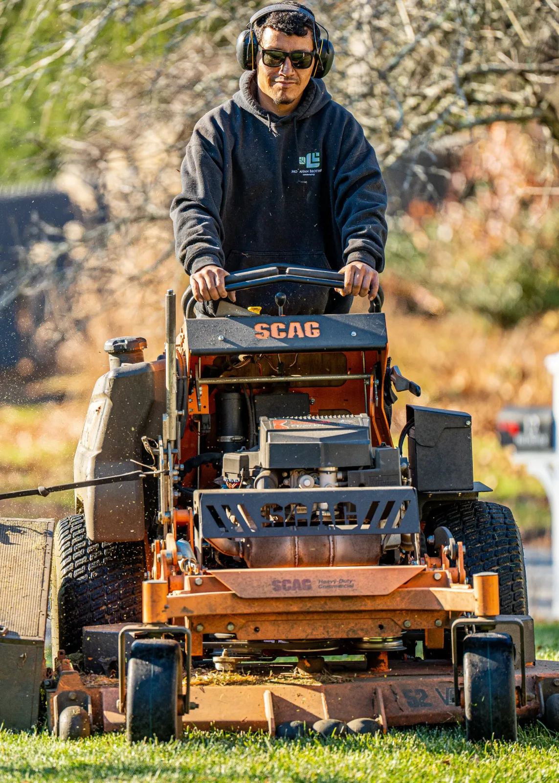 A man operating a large, orange SCAG lawn mower in a yard, wearing black headphones, sunglasses, and a black hoodie with a logo.