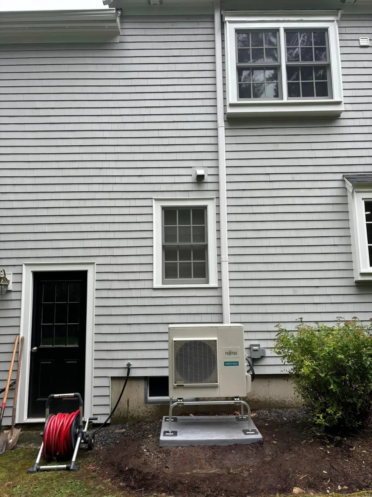 Back of a gray house with white-trim windows, utility boxes, a hose reel, and shrubs by the wall