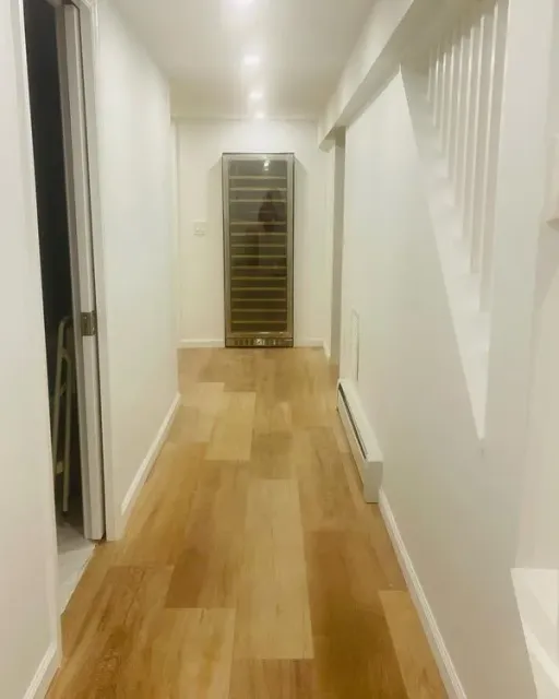 Narrow hallway with wood flooring, white walls, wine cooler, and stairs.