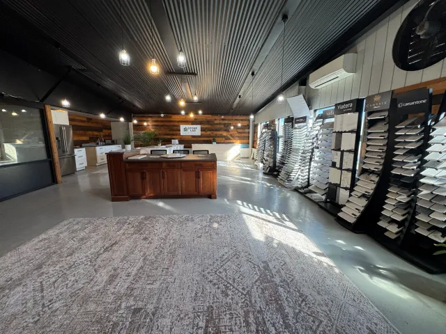 Interior of a flooring and tile showroom featuring display stands with samples, a central wooden island, seating area, and a wood-paneled wall with a sign that reads 'Stone Lab Surfaces,' illuminated by hanging lights.