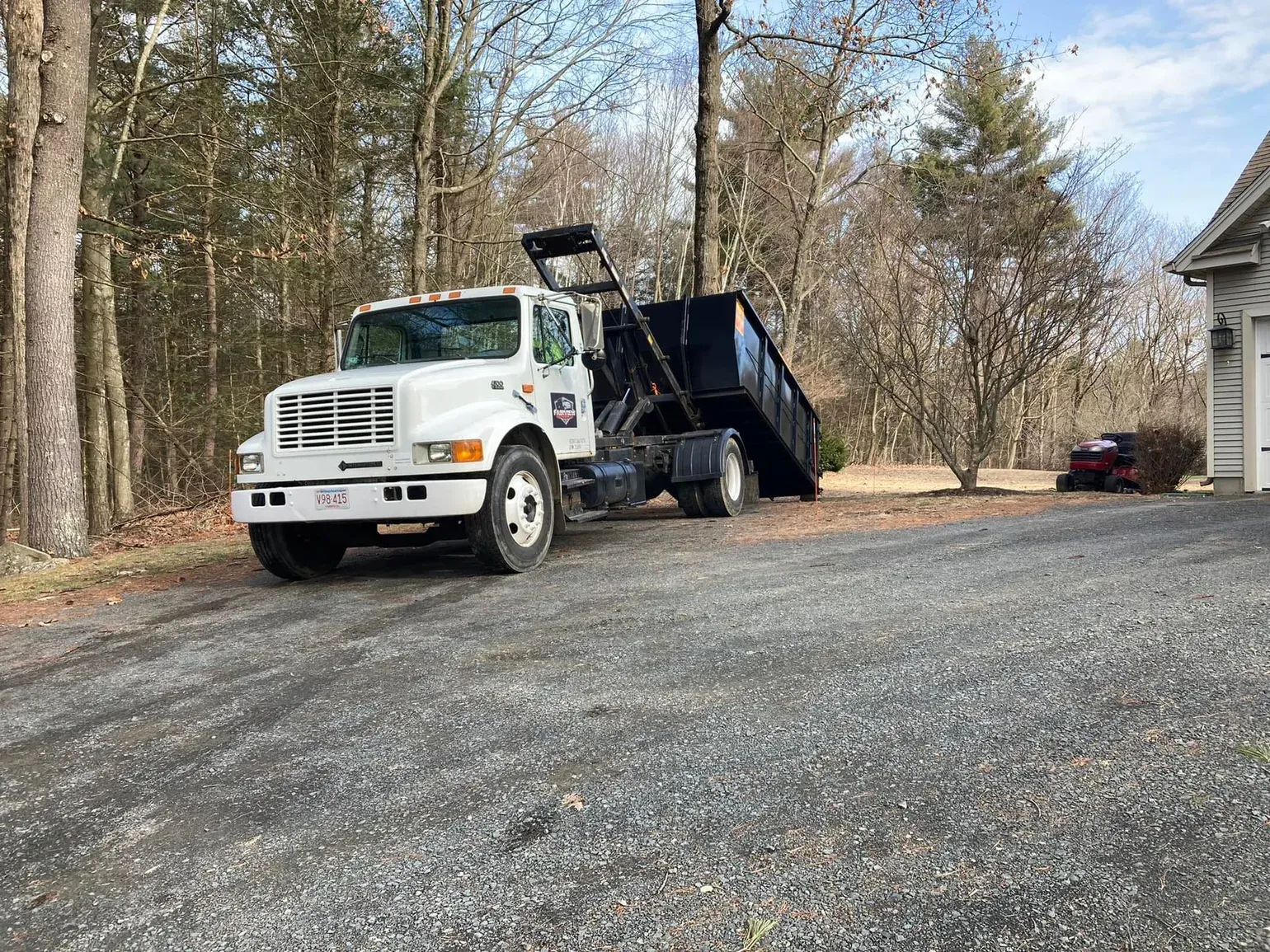 White dump truck with black container on a gravel driveway in front of a house.
