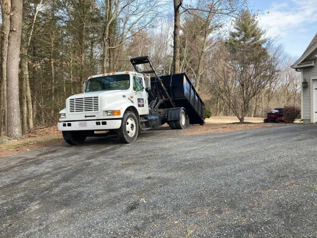 White dump truck with black container on a gravel driveway in front of a house.