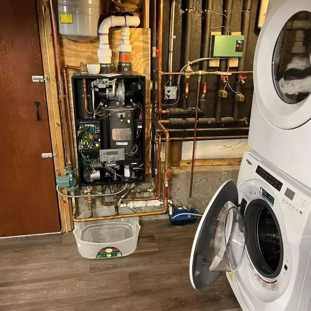 Laundry room with open washing machine and exposed boiler system above a bucket on the floor