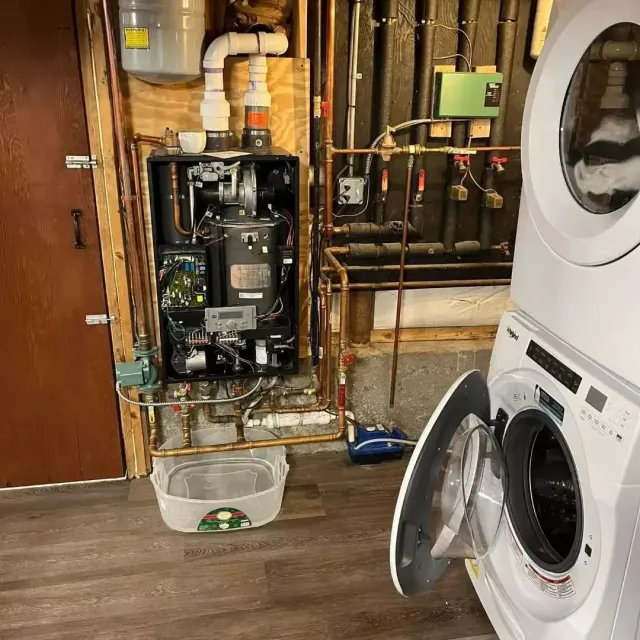 Laundry room with open washing machine and exposed boiler system above a bucket on the floor