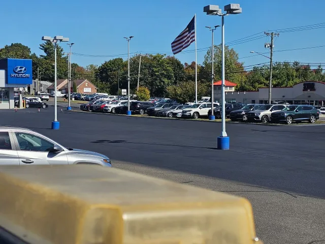 An outdoor Hyundai car dealership lot under a clear blue sky, featuring rows of parked vehicles and a waving US flag.