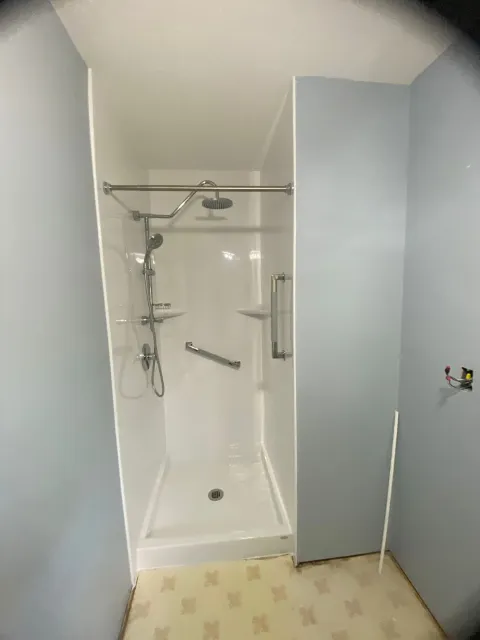 Empty shower with two shower heads, a grab bar, and soap shelves, with light blue walls and patterned linoleum flooring.