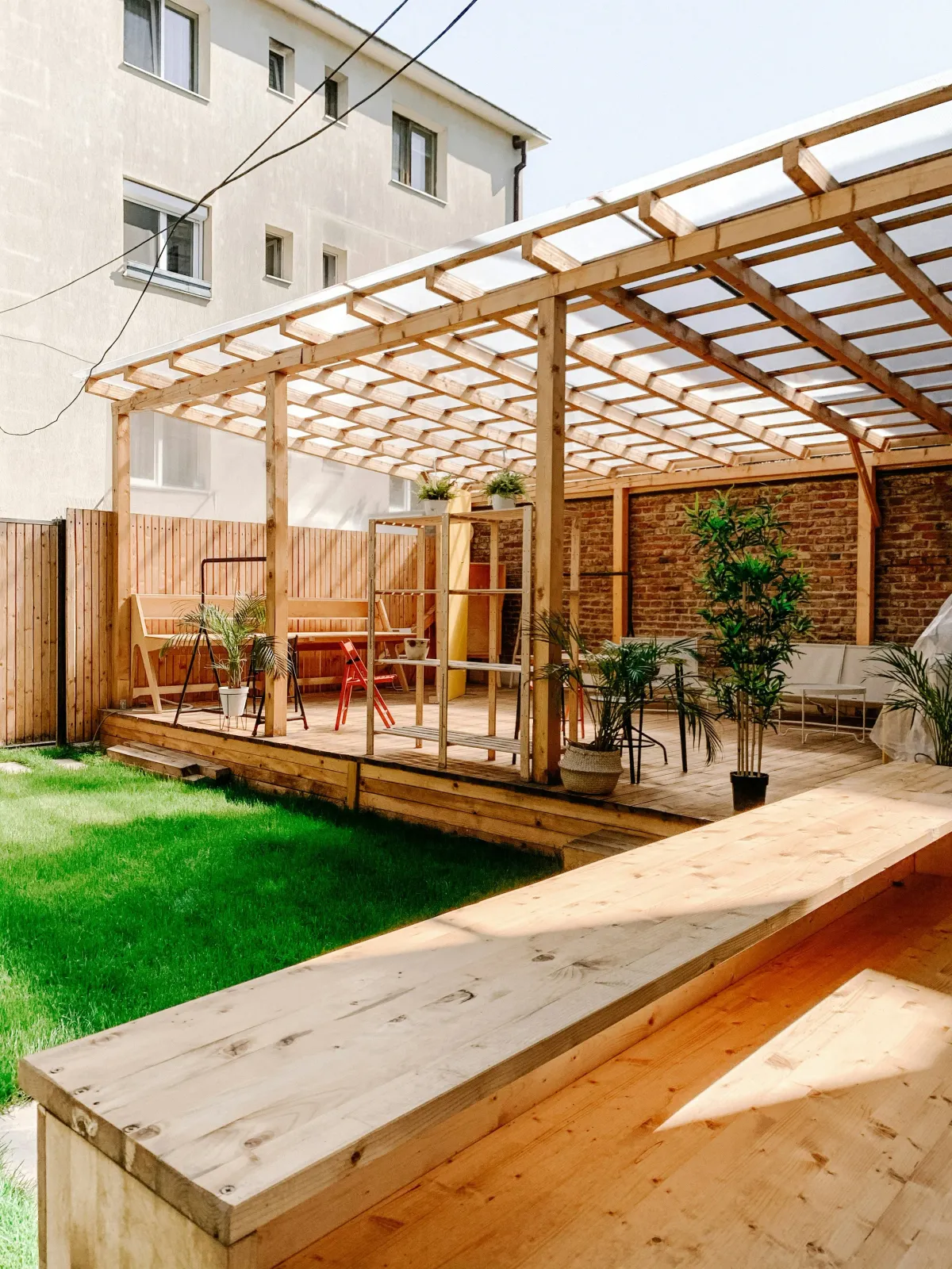 Backyard with a wooden patio and a pergola, surrounded by a wooden fence, with potted plants, outdoor furniture, and a house with multiple windows in the background.