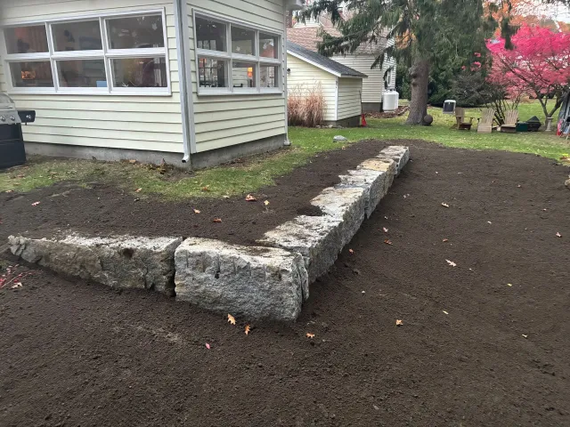 A stone retaining wall in a yard, near a light-colored house. Brown soil surrounds the wall and grass.