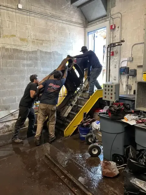 Three workers maneuver a large dark object up a short set of yellow industrial stairs inside a workshop.