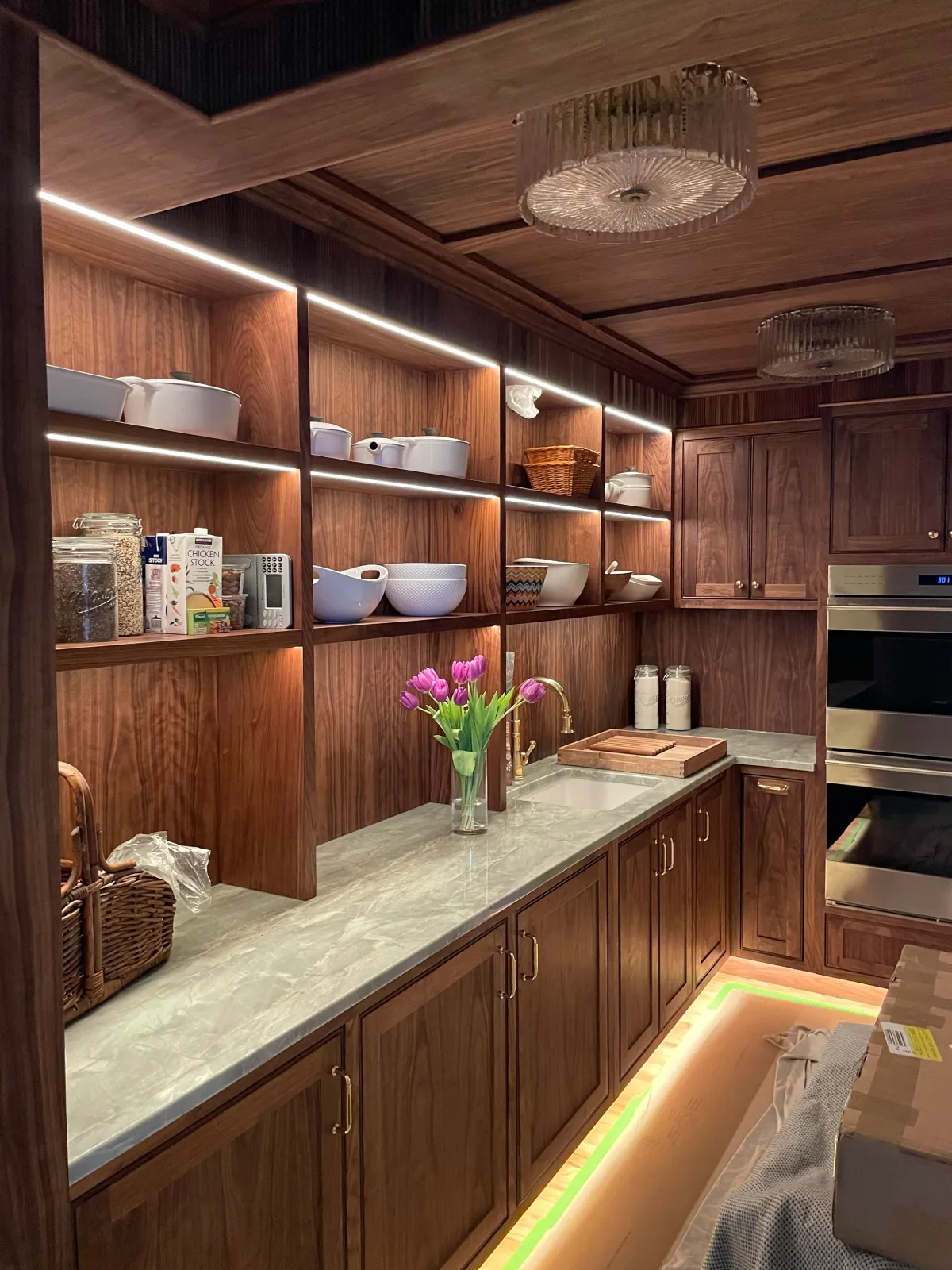 Kitchen with wooden cabinets, open shelving with dishes, a marble countertop, a vase with pink tulips, and built-in oven.