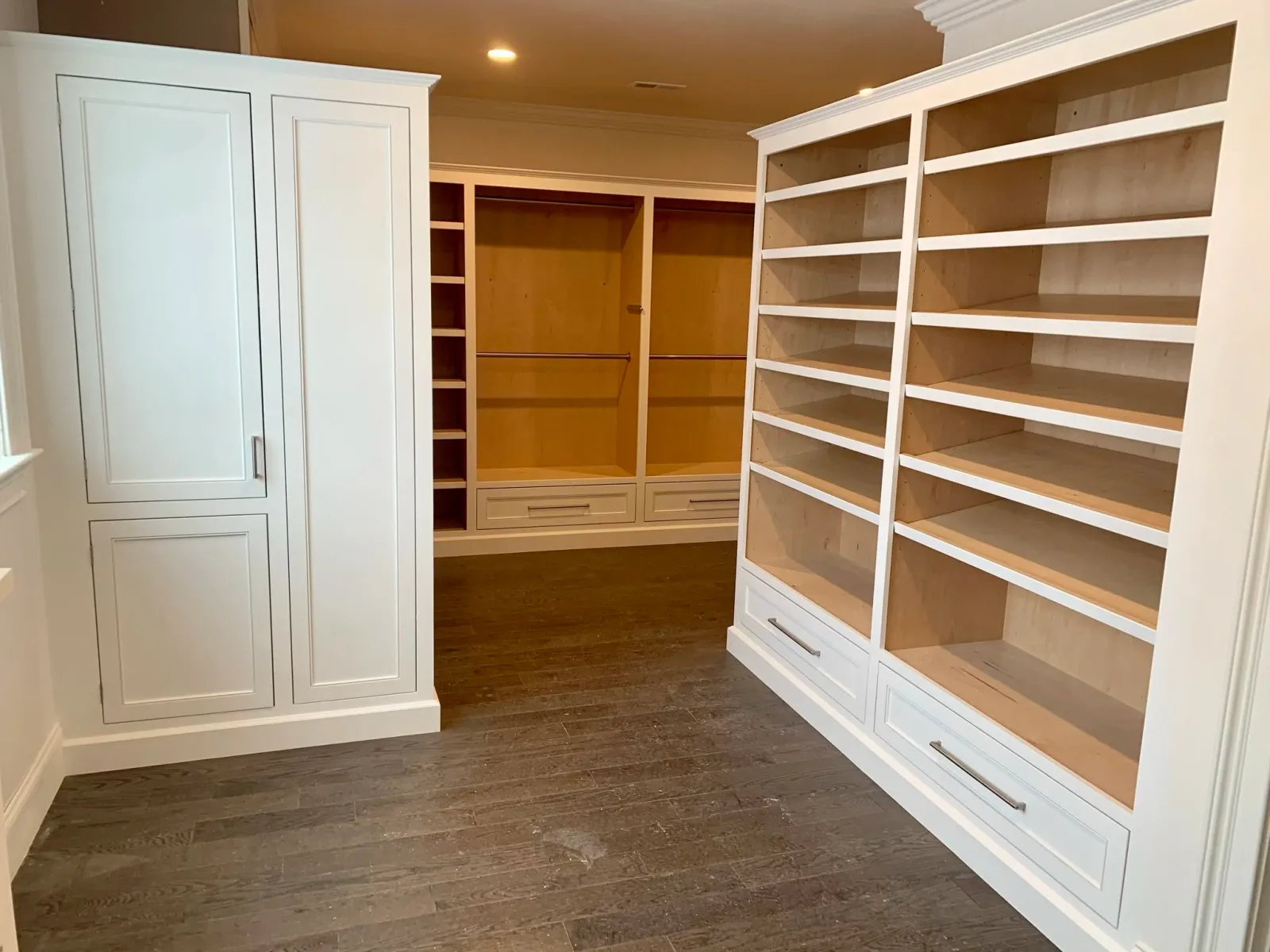 Empty walk-in closet with built-in white shelves and cabinets, with dark wooden flooring and overhead lighting.