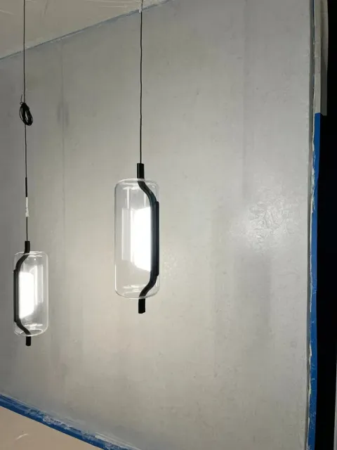 Two modern pendant lights with cylindrical glass shades and black hardware hanging against a plain light gray wall.