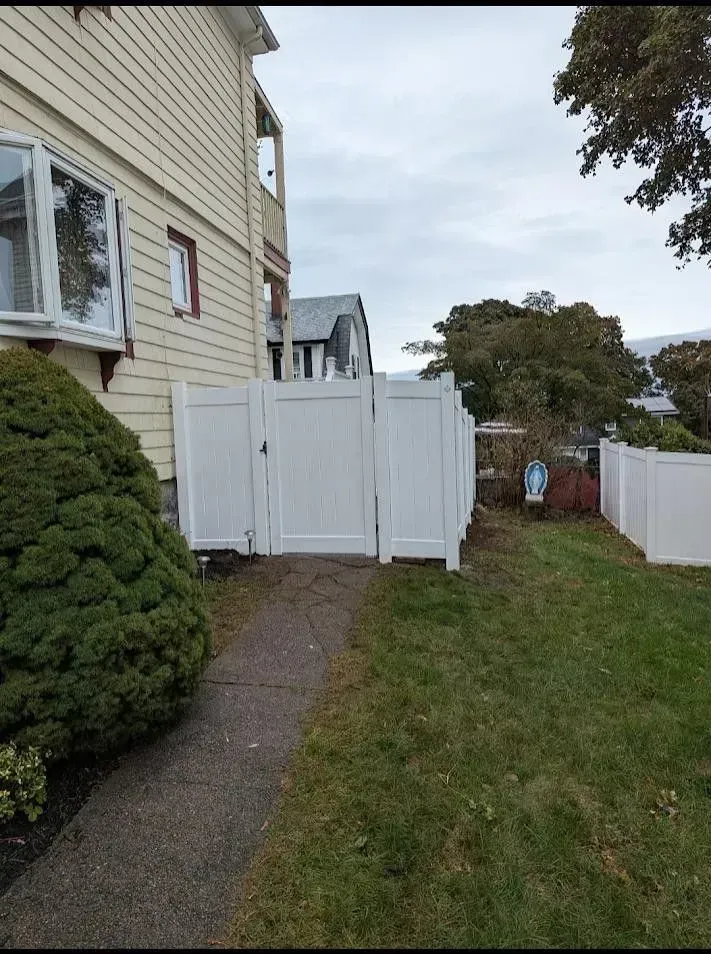 White backyard gate beside a beige house, opening to a grassy yard and paved side path under an overcast sky