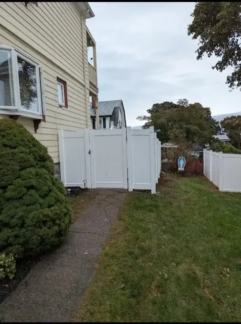 White backyard gate beside a beige house, opening to a grassy yard and paved side path under an overcast sky