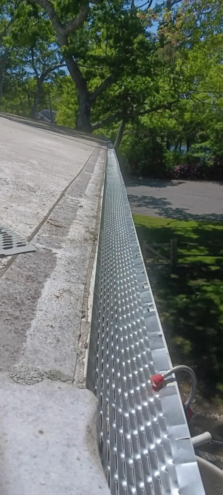 Sloped shingle roof beside a silver chain-link fence, with trees and a road below.