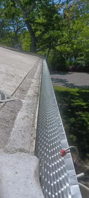 Sloped shingle roof beside a silver chain-link fence, with trees and a road below.