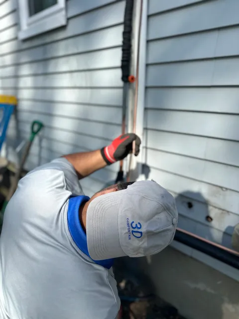 A person wearing a light gray cap, gray work shirt, and gloves is working on a water pipe or electrical conduit on the side of a house with light blue vinyl siding.