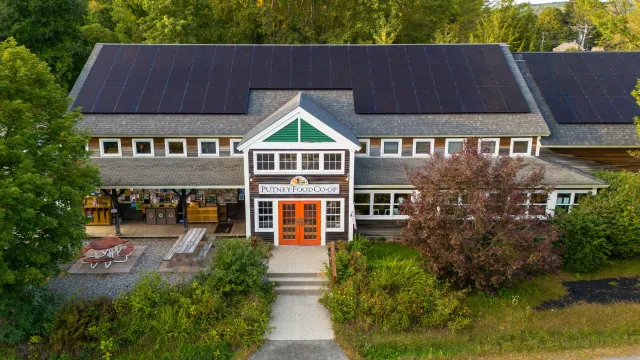 Aerial view of a building with a sign that reads 'Putney Food Co-op'. The building has a gray roof with two large solar panels, a front porch with a picnic table, and is surrounded by trees and greenery.
