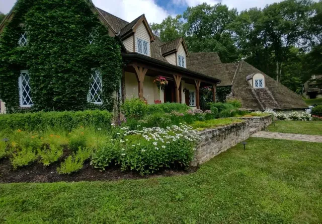 Cottage with a thatched roof, covered in ivy. A stone wall borders a garden of flowers and green shrubs.