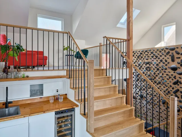 Interior view of a modern home staircase with wooden steps and black metal railings, leading to an upper loft area with a red sofa, green chair, and large windows.
