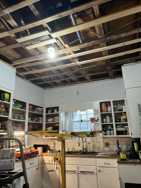 A partially remodeled kitchen with exposed ceiling beams, a bright light bulb, and open cabinets filled with dishes and kitchen items. There's a window above the sink and some tools and equipment on the counter and floor.