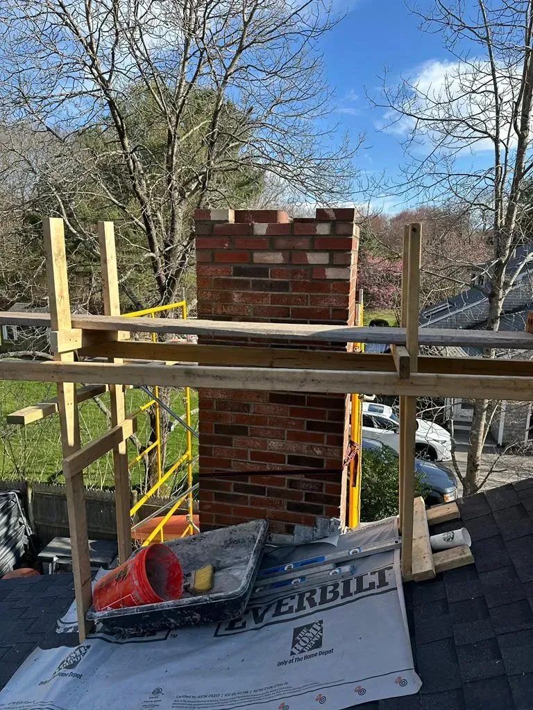 Brick chimney under construction on wooden scaffolding with tarp and trees in the background