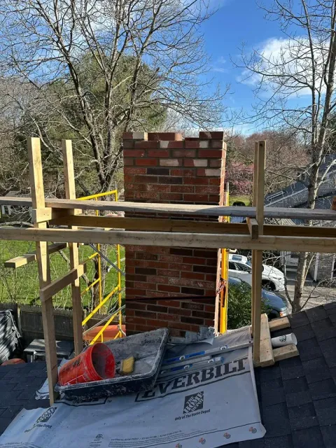 Brick chimney under construction on wooden scaffolding with tarp and trees in the background