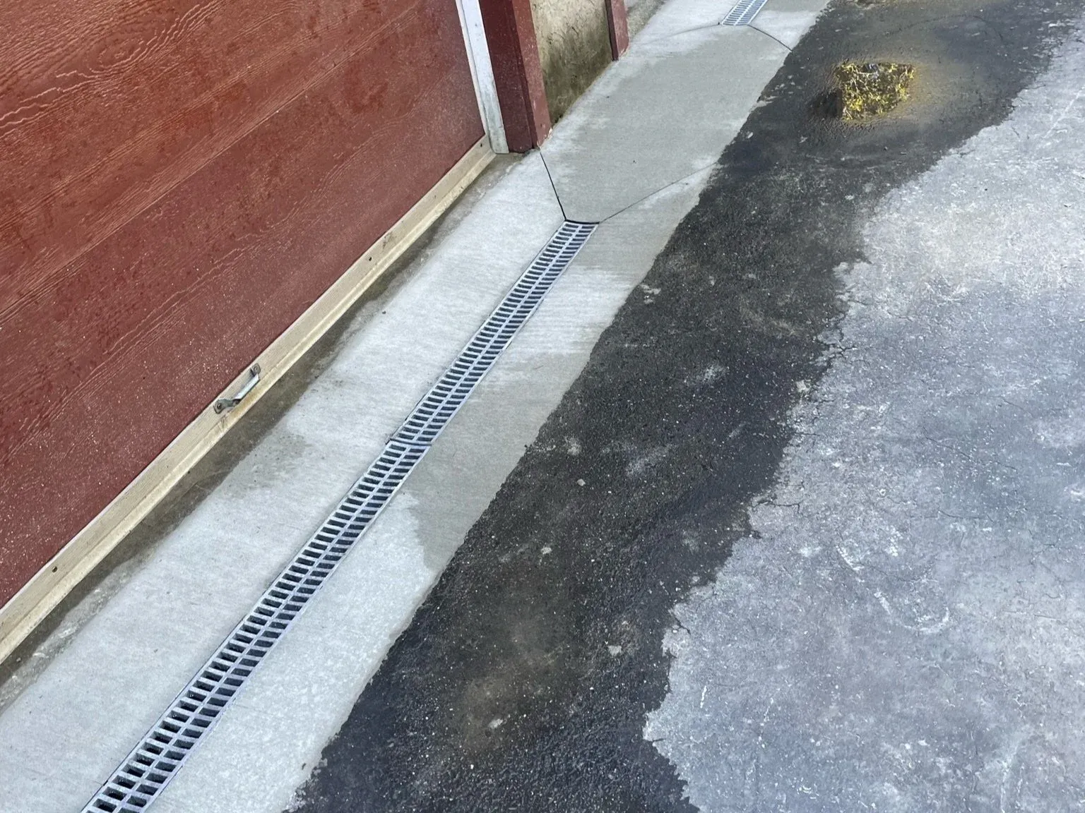 Drainage grate along concrete sidewalk beside red wooden building, with wet and dry asphalt pavement.
