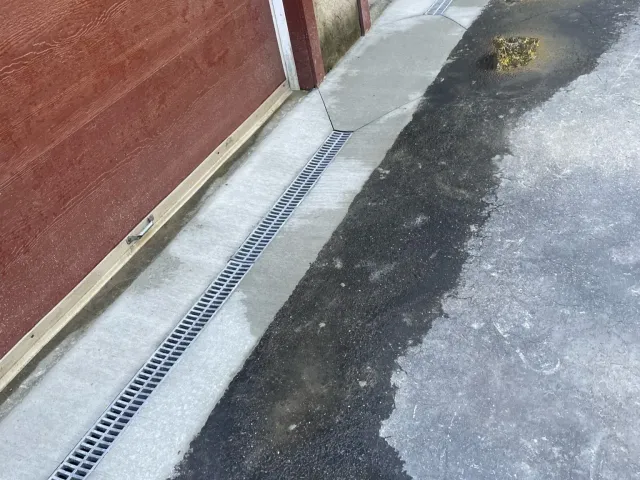 Drainage grate along concrete sidewalk beside red wooden building, with wet and dry asphalt pavement.