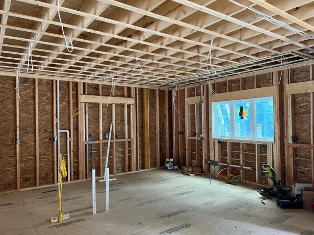 An interior room under construction with wood wall framing, a grid ceiling, exposed pipes, and a window with blue film.