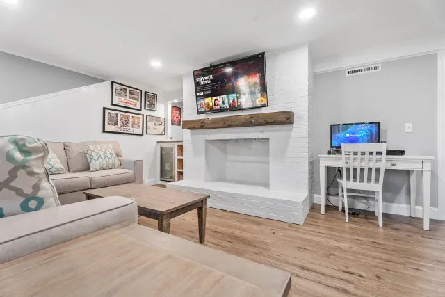 Modern living room with white painted brick fireplace, wall-mounted TV displaying Netflix 'Stranger Things,' beige sofa with patterned pillows, small wooden coffee table, wall art, and a desk with a computer showing Windows 11, all on hardwood floors with white walls and ceiling lights.