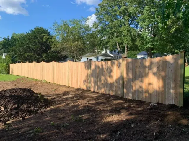 A new, light wood privacy fence with a scalloped top edge borders a dirt patch in a residential yard.
