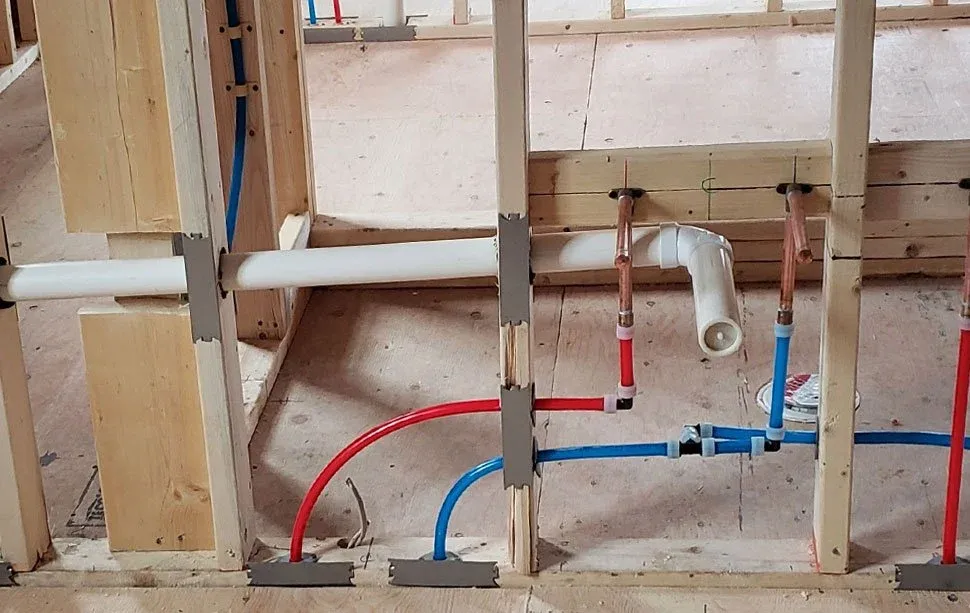 Construction framing with visible white, red, and blue plumbing pipes installed on a wooden wall frame.