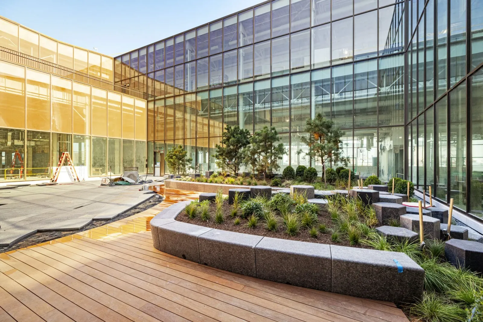 Modern outdoor courtyard in Boston's Seaport District under construction with glass building, wooden decking, and landscaped garden with small trees and shrubs.