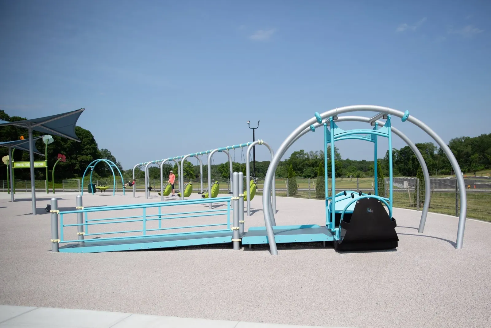 Empty outdoor playground with metal swings, slides, and climbing structures under a blue sky, surrounded by trees.