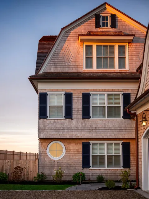 A three-story house with beige shingles, black shutters, and a gray roof. The house features a circular window on the first floor, large double-pane windows with shutters on the second and third floors. The house is illuminated by warm sunset light, with a small garden and a wooden fence in the yard.
