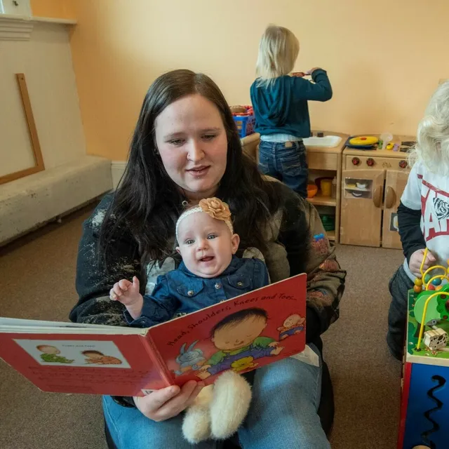 A woman reads to a smiling baby while children play in the background.