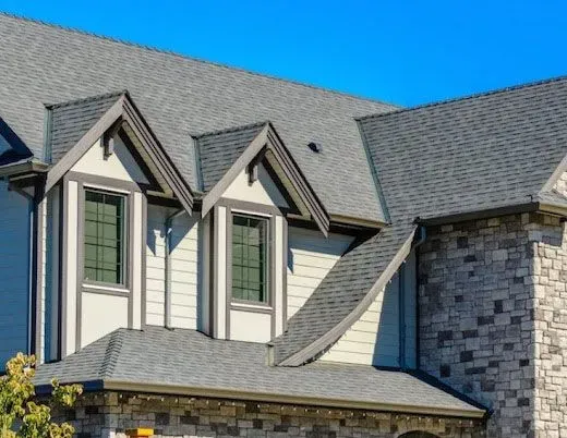 Stone and white house exterior with gray shingled roof and dormer windows under a blue sky