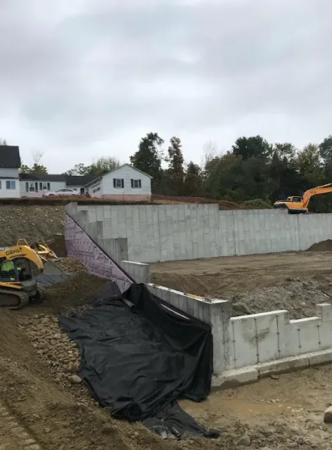 Construction site with concrete retaining wall, excavator, and black tarp on an embankment under cloudy sky