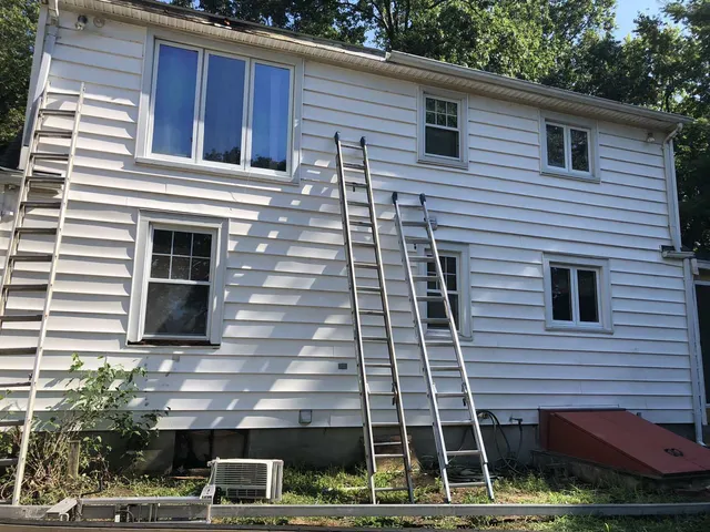 A two-story white house with siding, three windows, and two extension ladders leaning against the exterior wall.