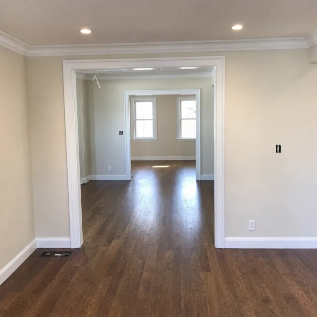 Empty beige room with hardwood floors, wide doorway, and two windows in the back.