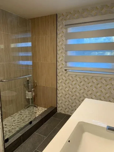 Bathroom with walk-in shower with glass doors, beige and patterned wall tiles, a white countertop with a sink, and a window with horizontal blinds.