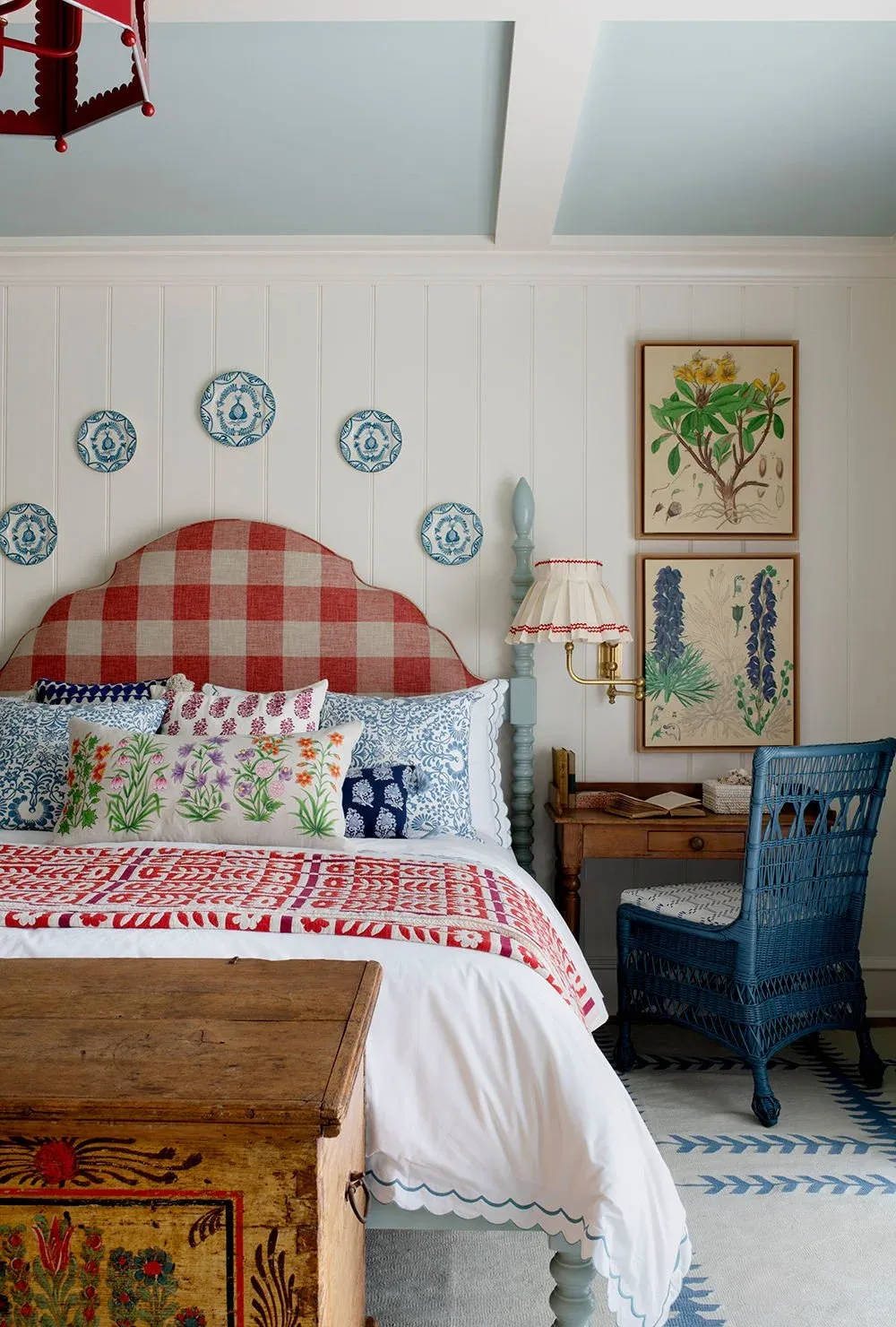 Cozy bedroom with a red checkered headboard, blue and white patterned pillows, botanical wall art, and a rustic wooden chest. Features a wicker chair and desk, a vintage lamp, and decorative plates on the walls.