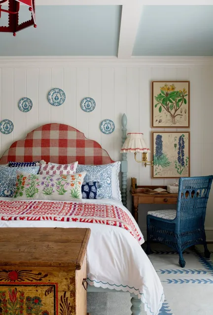 Cozy bedroom with a red checkered headboard, blue and white patterned pillows, botanical wall art, and a rustic wooden chest. Features a wicker chair and desk, a vintage lamp, and decorative plates on the walls.