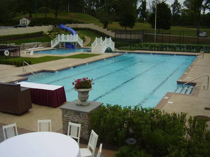 A swimming pool with a water slide and a small water play structure. There are lounge chairs, a table with chairs, and decorative plants surrounding the pool area.